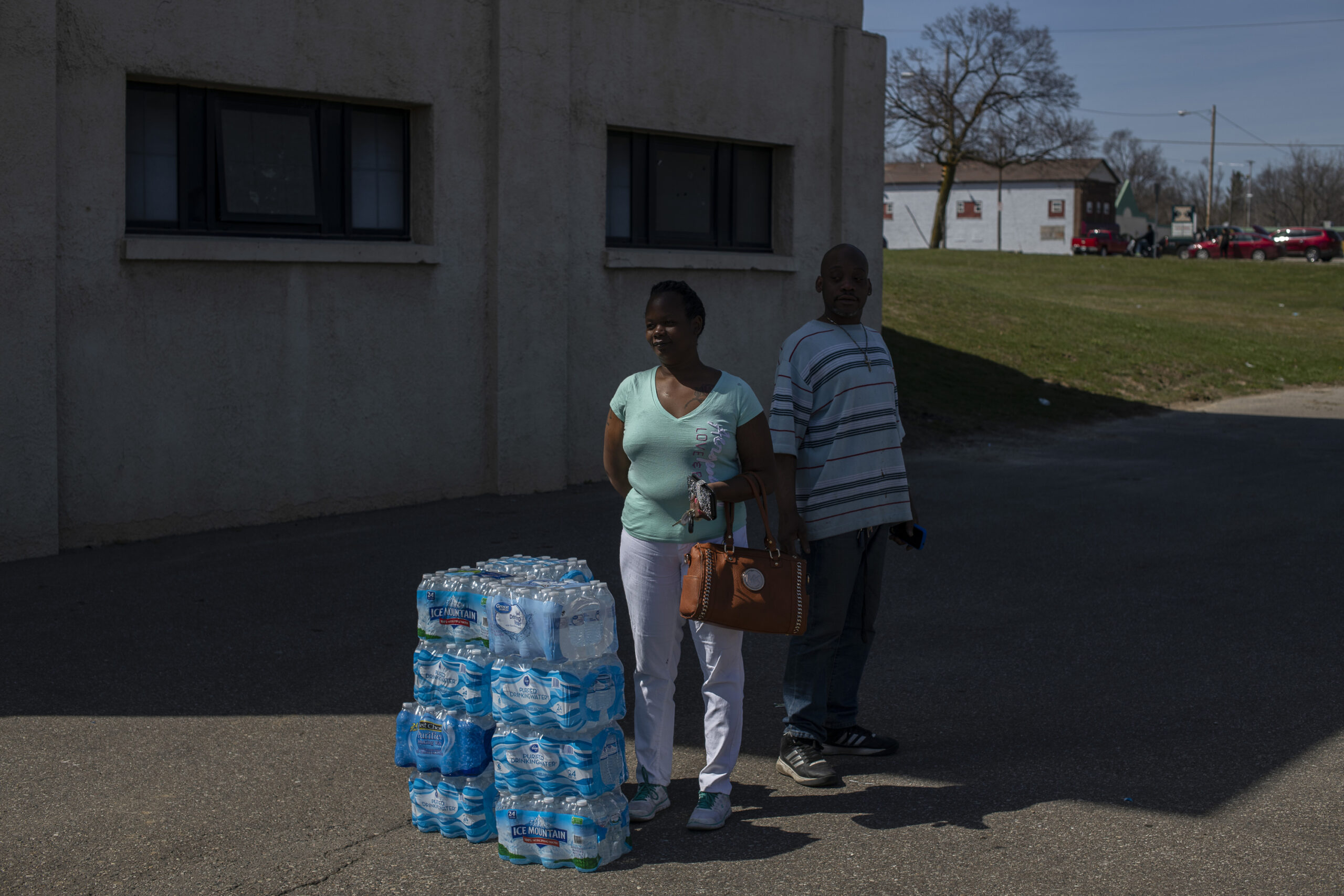 A Black woman and man stand next to a stack of packages of bottled water on the pavement in the shadow of a concrete building.