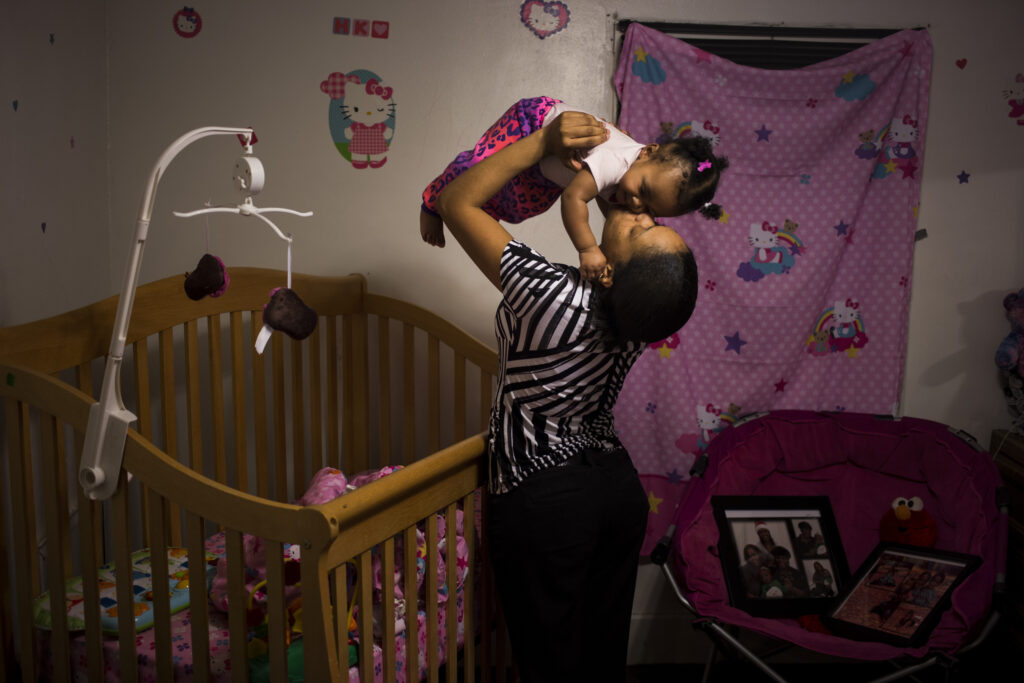 A Black woman kisses her baby on the cheek while lifting her in the air above her crib. The baby’s room is decorated in a Hello Kitty theme. 