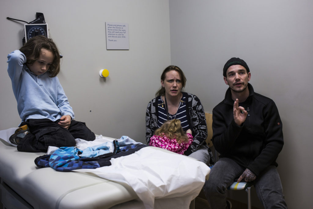 A white man and woman look concerned as they address their son’s doctor (not pictured) in a medical examination room. The son sits on his knees on the examination table. 