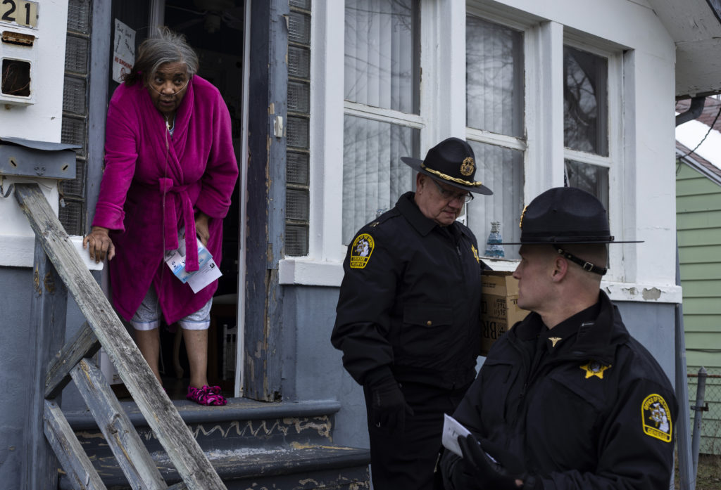 An older Black woman opens the door to her home to greet two white male police officers. One holds a cardboard box (of water filters.) 