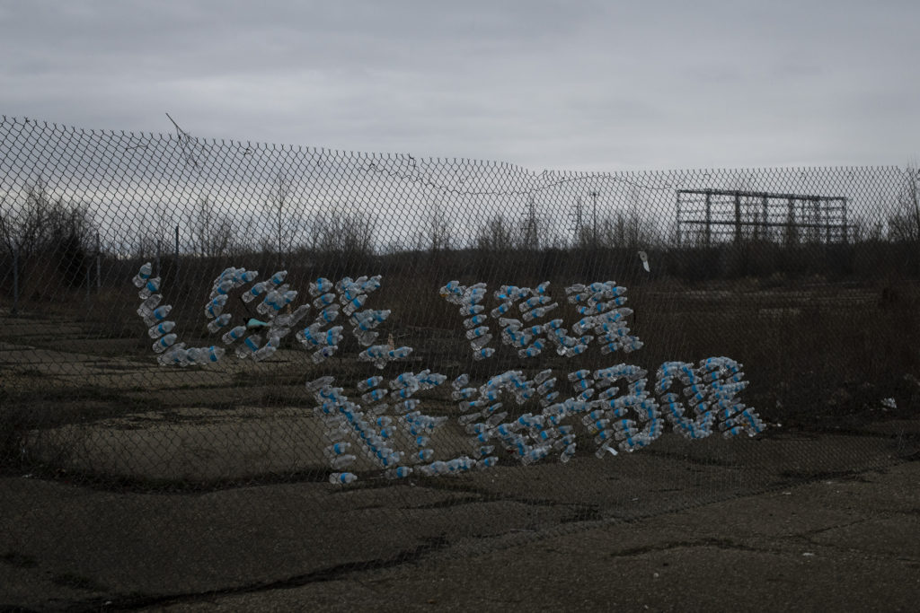The phrase “love your neighbor” is spelled out in empty water bottles shoved through the holes of a chain link fence. 