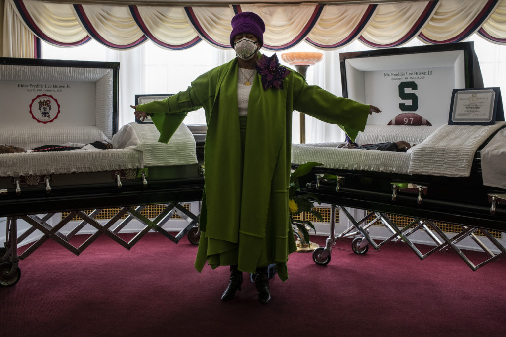 A Black woman in a green dress with a large purple broach and a purple hat stands with her arms outstretched between two caskets in the parlor of a funeral home. The casket to her left holds an older Black man, and the casket to the right holds a younger Black man. Both caskets are lined with white silk. 