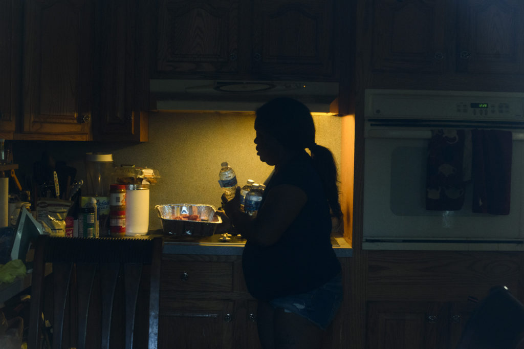 A Black woman stands in silhouette in her kitchen while holding several bottles of water. 