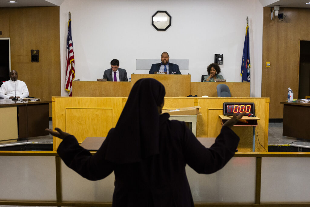 A woman stands with her hands raised as she speaks to three City Council members sitting at raised, conjoined desks in front of her. 
