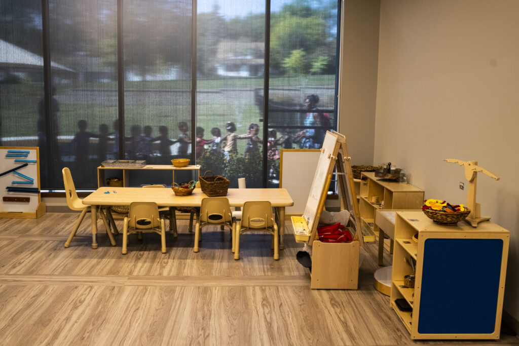 Small children form a single-file line outside of the window of an empty children’s classroom. 
