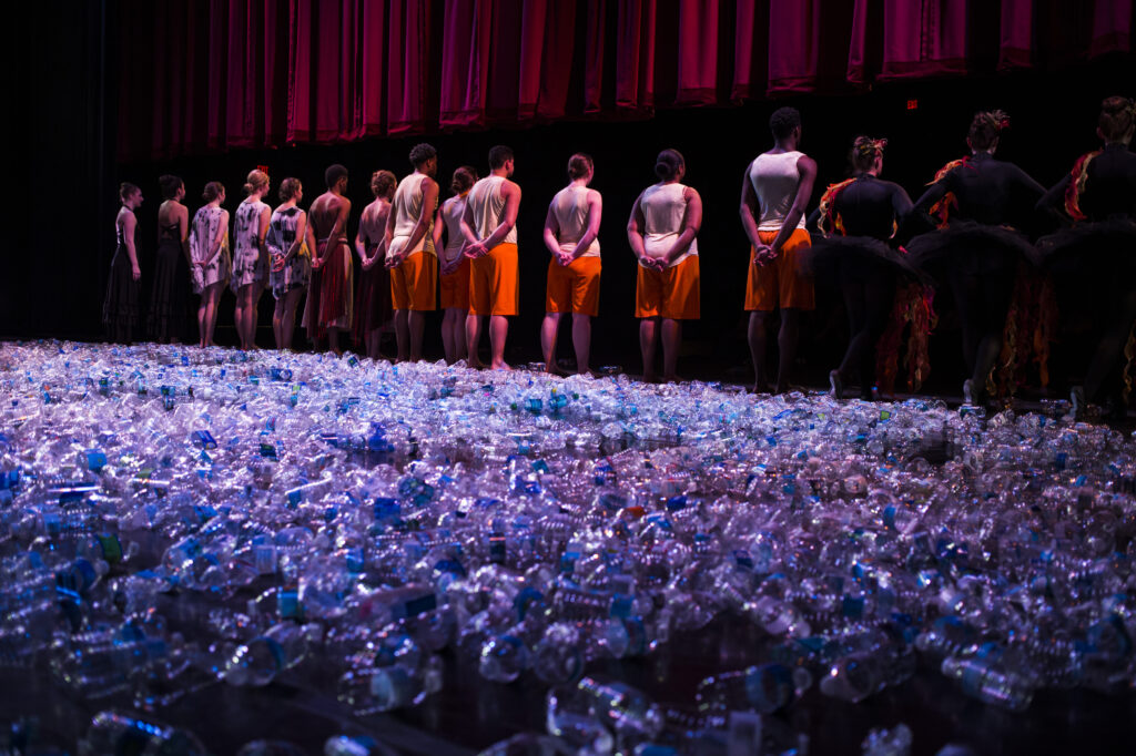 Sixteen dancers form a line at the front of a stage. Hundreds of empty water bottles blanket the stage behind them. 
