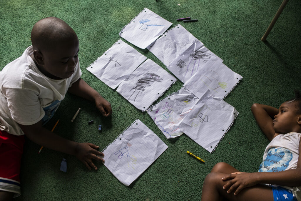 Two small Black children lay together on a bright green carpet. Loose leaf papers with their drawings are strewn on the ground between their bodies. 
