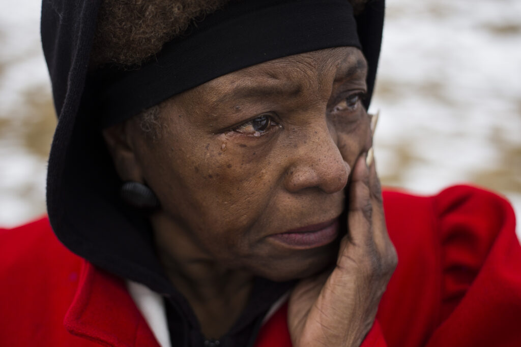 An older Black woman wipes tears from her face while standing outside. A black hood covers her head, and she wears a bright red wool coat. 
