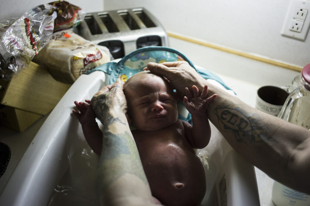 A white man with tattooed arms washes a newborn baby in a basin on a kitchen counter. 

