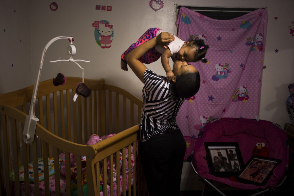 A Black woman kisses her baby on the cheek while lifting her in the air above her crib. The baby’s room is decorated in a Hello Kitty theme. 
