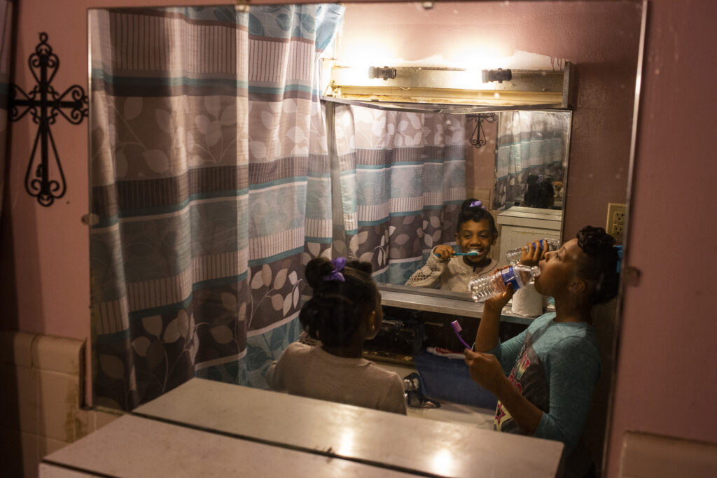 Two Black girls brush their teeth in a pink bathroom. They rinse their mouths with bottled water. 
