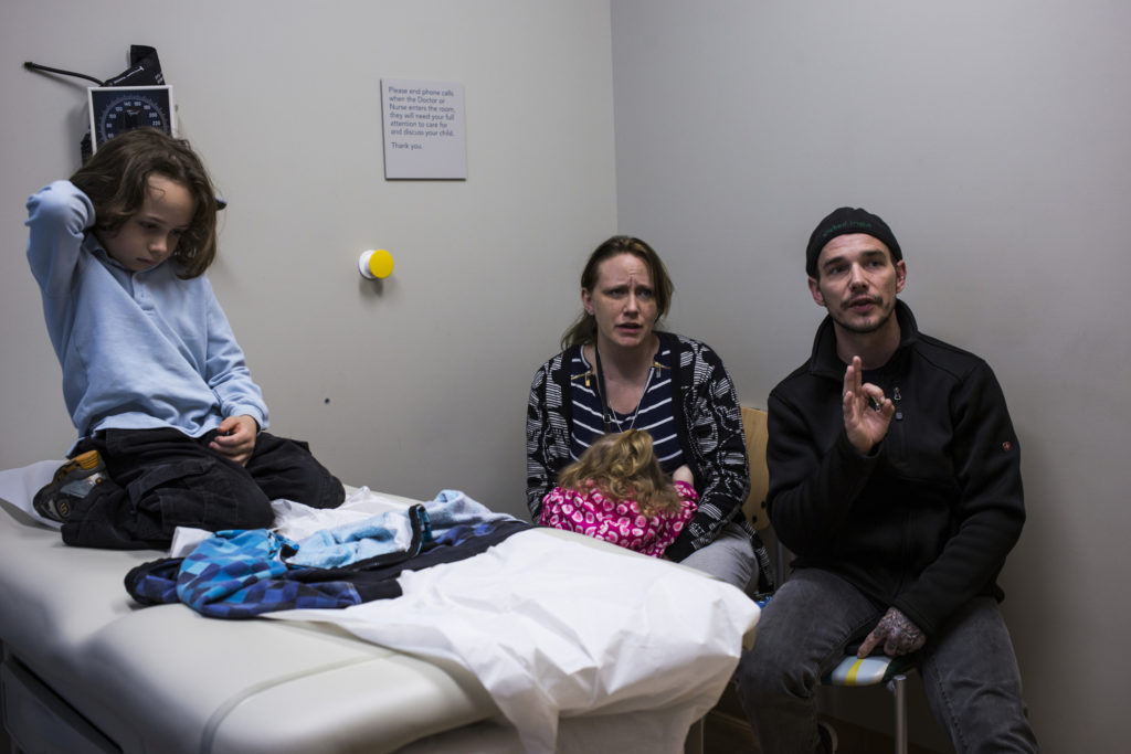 A white man and woman look concerned as they address their son’s doctor (not pictured) in a medical examination room. The son sits on his knees on the examination table. 
