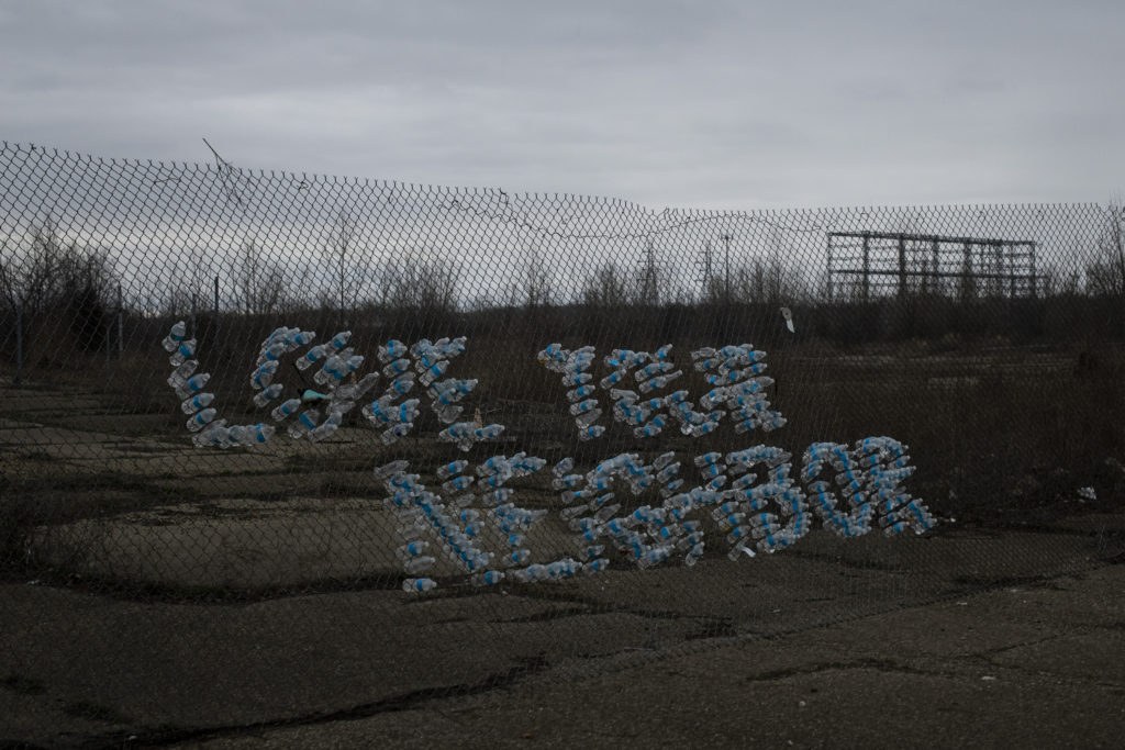 The phrase “love your neighbor” is spelled out in empty water bottles shoved through the holes of a chain link fence. 
