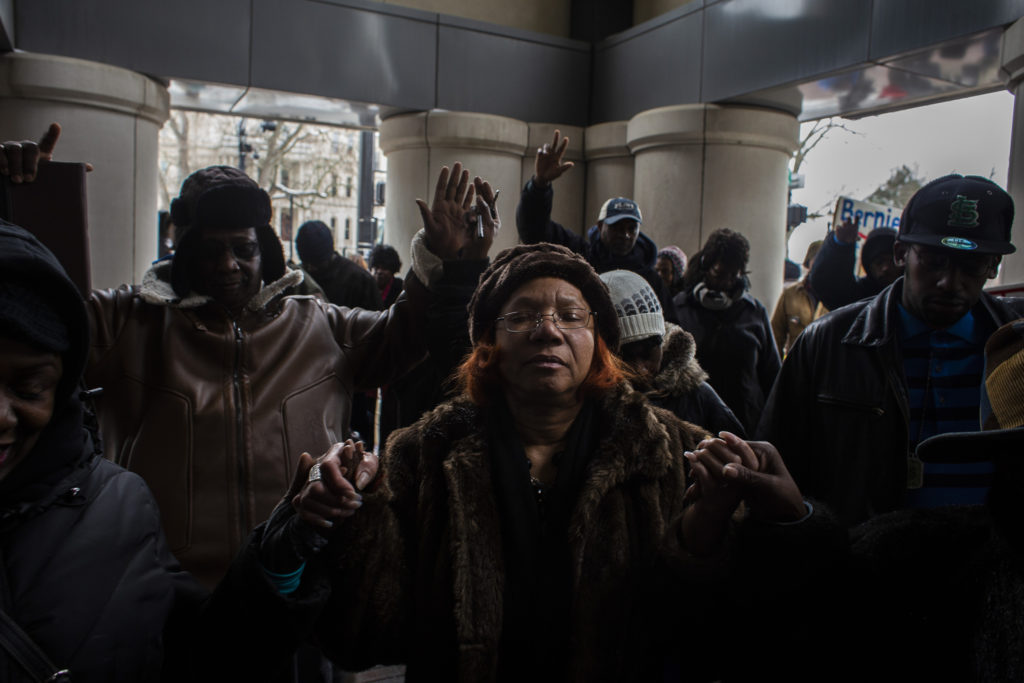 A Black woman with red hair wearing a brown fur coat joins hands with other Black protesters outside of a building. 
