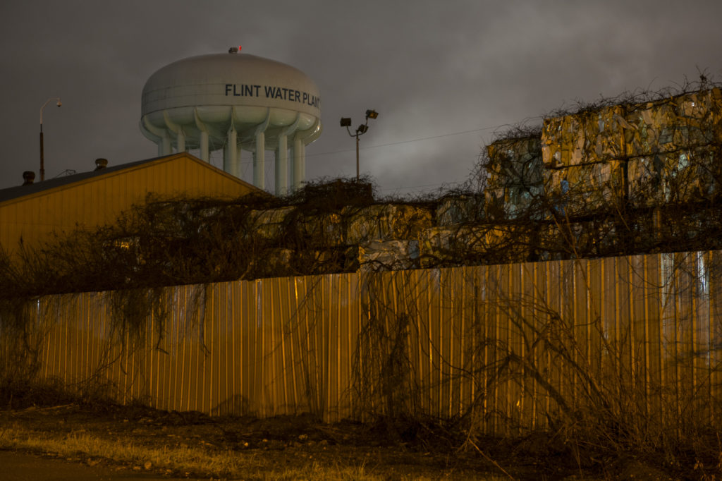 A water tower that says “Flint Water Plant” rises behind a metal scrap yard. 
