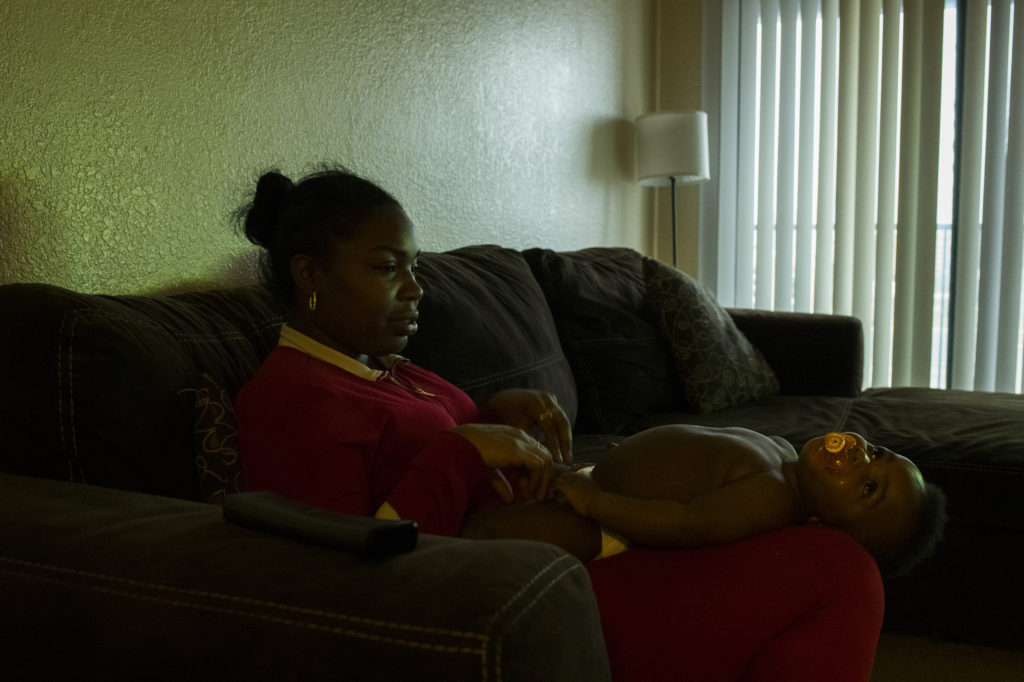 A Black woman in a red outfit sits on the couch while her son reclines in her lap. 
