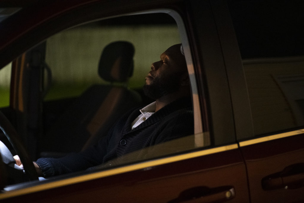 A Black man closes his eyes as he rests in the front seat of his car. 
