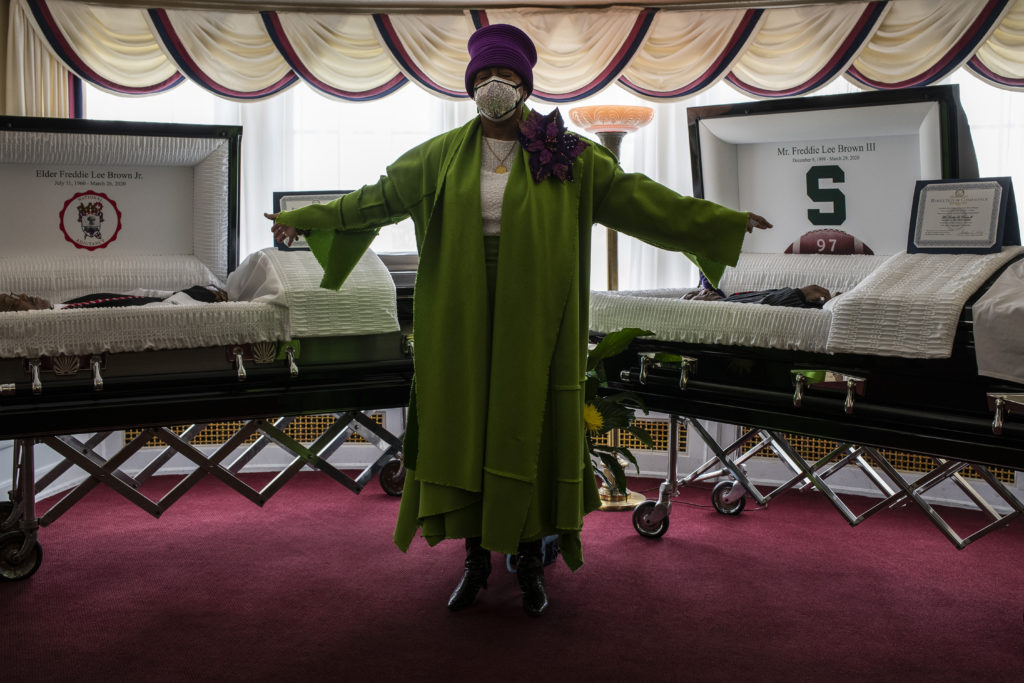 A Black woman in a green dress with a large purple broach and a purple hat stands with her arms outstretched between two caskets in the parlor of a funeral home. The casket to her left holds an older Black man, and the casket to the right holds a younger Black man. Both caskets are lined with white silk. 
