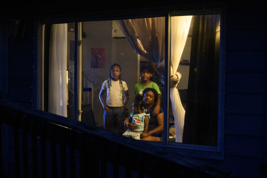 A Black woman with two young children standing behind her and a baby in her lap are are photographed through their living room window. 
