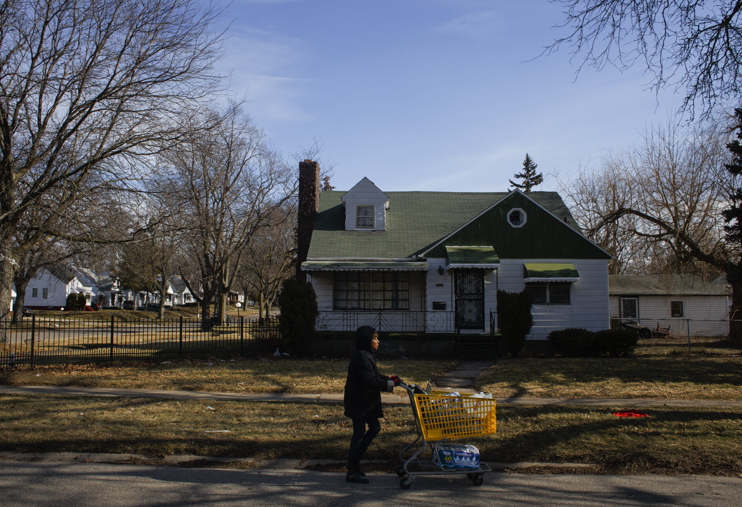 Person pushing orange shopping cart in foreground, white and green house in background