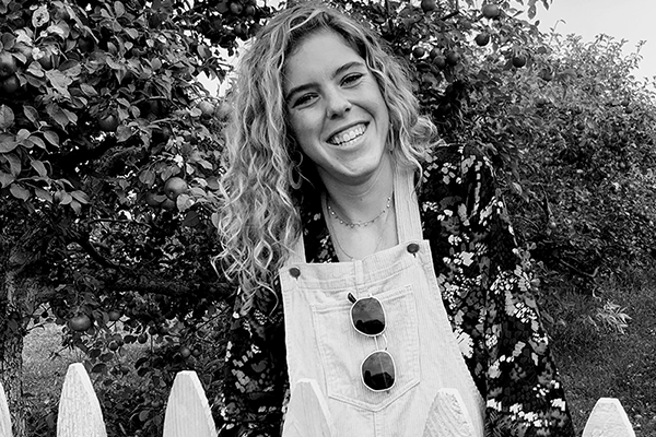Brigid smiles next to a picket fence while wearing a patterned shirt and light colored overalls