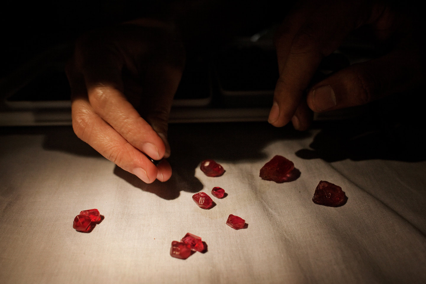 Two hands inspect red spinel gemstones on a cloth covered table.