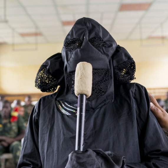 A person holds a microphone and wears a black robe, black gloves, and a black mask covering their entire face