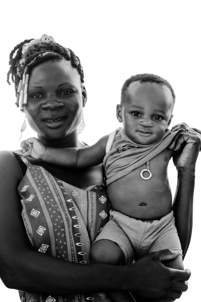 A woman holds a small child, both looking at the camera, with a white background behind them