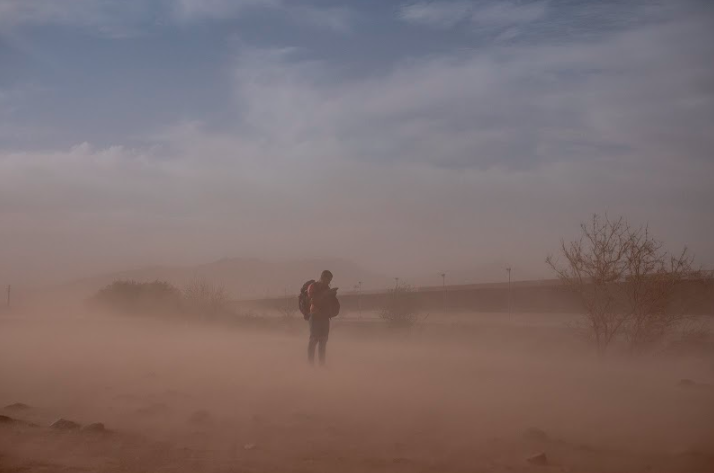 A person on-the-move checks their phone in the middle of a dust storm near Door 36, an
official service gate operated by Border Control, of the fence along the U.S. border
separating El Paso, Texas, from Ciudad Juárez. Photo by Oscar B. Castillo and Wil Sands.